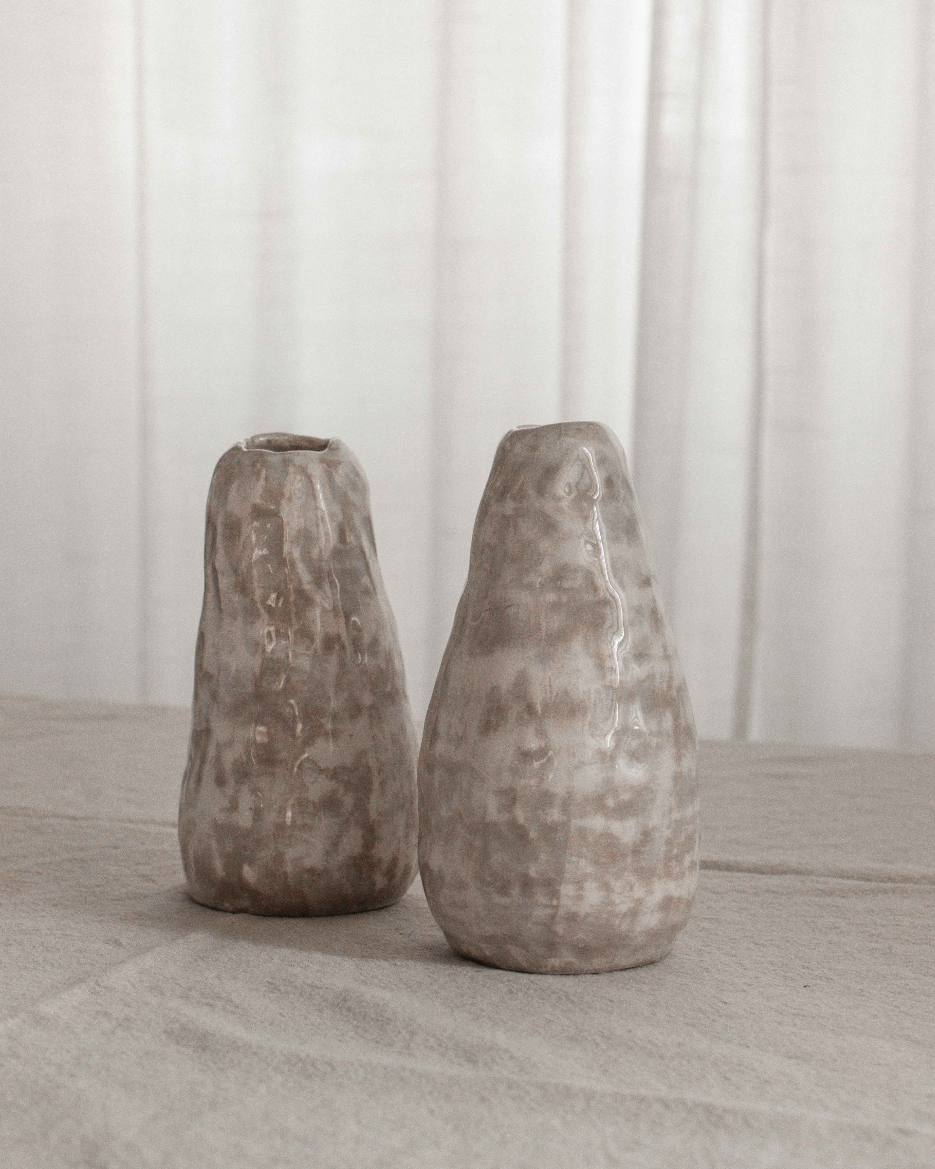 Two textured stone vases on a light wooden surface with a white curtain background.
